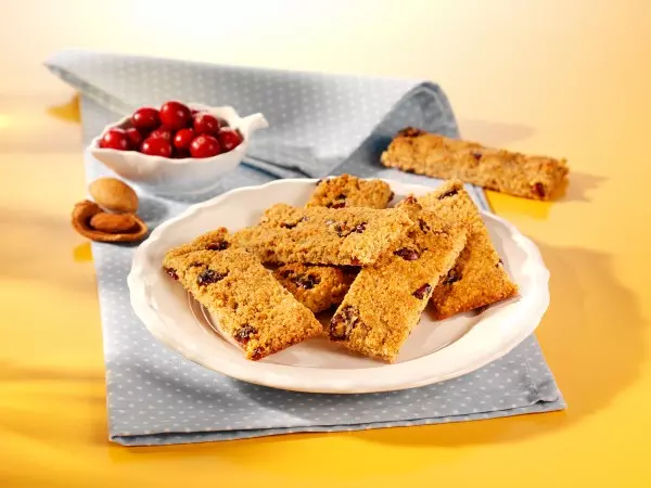 MycoVital_ Un piatto bianco con barrette d'avena rettangolari contenenti frutta rossa, appoggiato su un panno blu a pois. Sullo sfondo, una piccola ciotola bianca di mirtilli rossi e alcune albicocche.