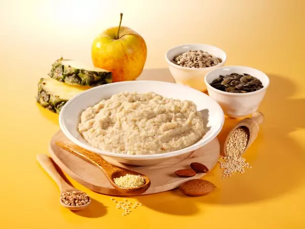 MycoVital_ Una ciotola di porridge cremoso è circondata da un cucchiaio di legno, fette di ananas, una mela, mandorle, semi di zucca, semi di girasole e misurini di cereali, il tutto su uno sfondo giallo.