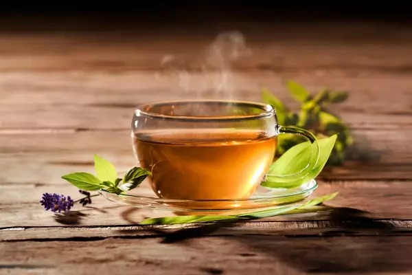 MycoVital_ Eine klare Glastasse, gefüllt mit dampfendem Kräutertee, steht auf einer passenden Untertasse, umgeben von frischen grünen Blättern und einer kleinen violetten Blume, alles auf einer rustikalen Holzfläche.