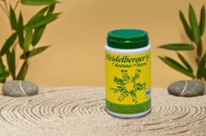 MycoVital_ Ein gr&uuml;ner und gelber Beh&auml;lter mit der Aufschrift Heidelberger's 7 Kr&auml;uter-Stern steht auf einer geharkten Sandfl&auml;che, umgeben von glatten Steinen und gr&uuml;nen Bl&auml;ttern vor einem beigen Hintergrund.