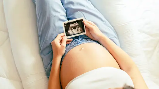MycoVital_ Eine schwangere Person liegt auf einem Bett und h&auml;lt ein Ultraschallbild &uuml;ber ihren Bauch, sie tr&auml;gt ein hellblaues Hemd und eine hellblaue Hose, von oben gesehen.