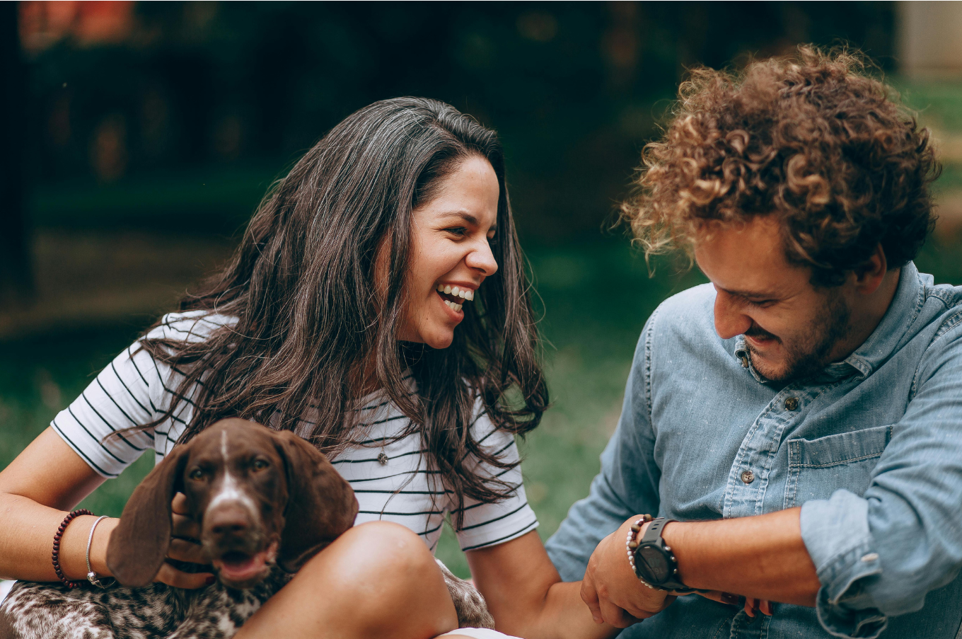 MycoVital_ Eine Frau und ein Mann sitzen eng beieinander im Freien, lächeln und lachen, während ein brauner Hund auf dem Schoß der Frau ruht. Die Gruppe wirkt glücklich und entspannt in einer grünen, grasbewachsenen Umgebung.
