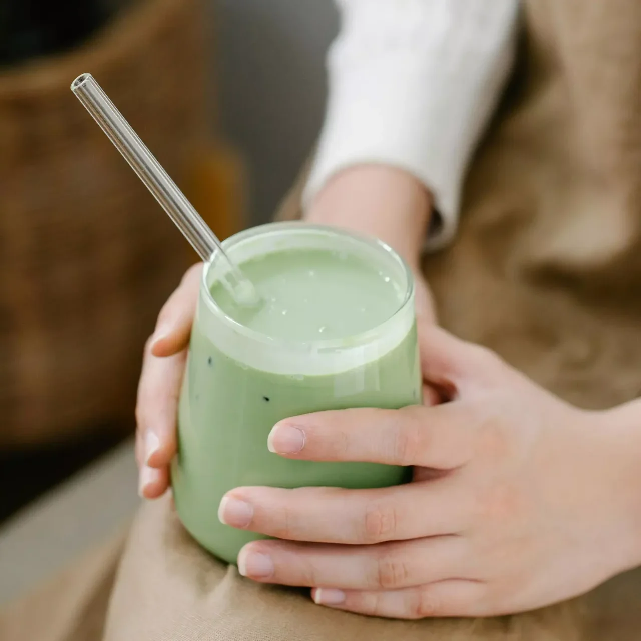 MycoVital_ Una persona che indossa una camicia leggera a maniche lunghe e un grembiule marrone tiene in mano un bicchiere di matcha latte verde con una cannuccia di vetro trasparente appoggiata sulle ginocchia.