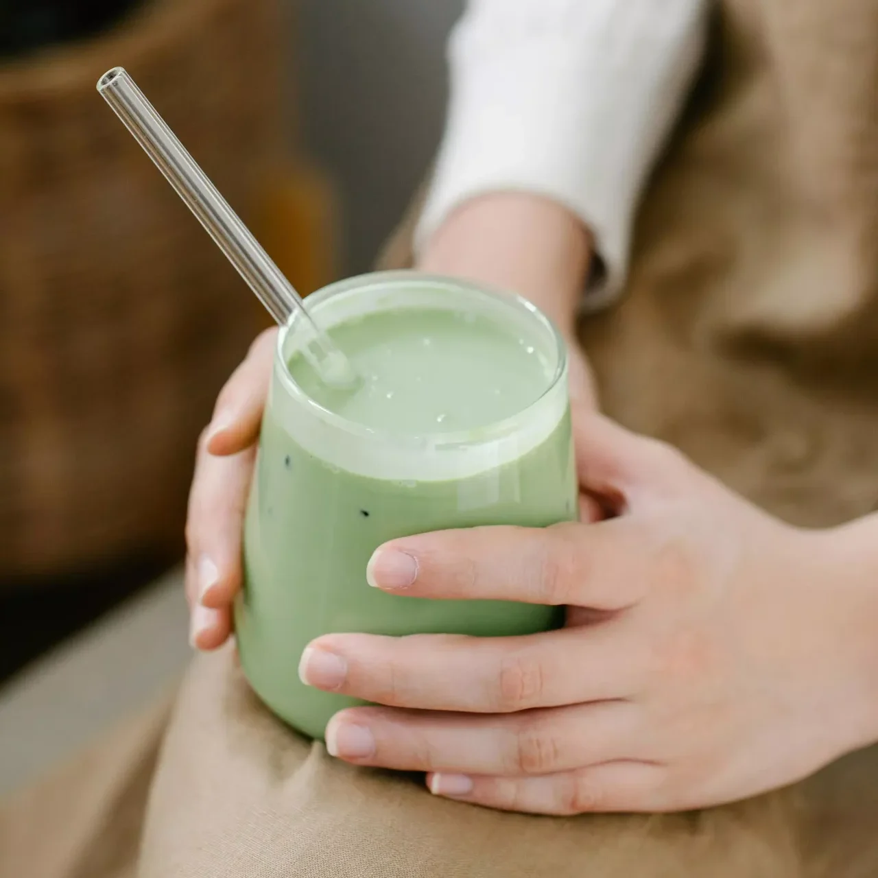 MycoVital_ Una persona con camisa clara de manga larga y delantal marrón sostiene un vaso de matcha latte verde con pajita de cristal transparente, apoyándolo sobre su regazo.