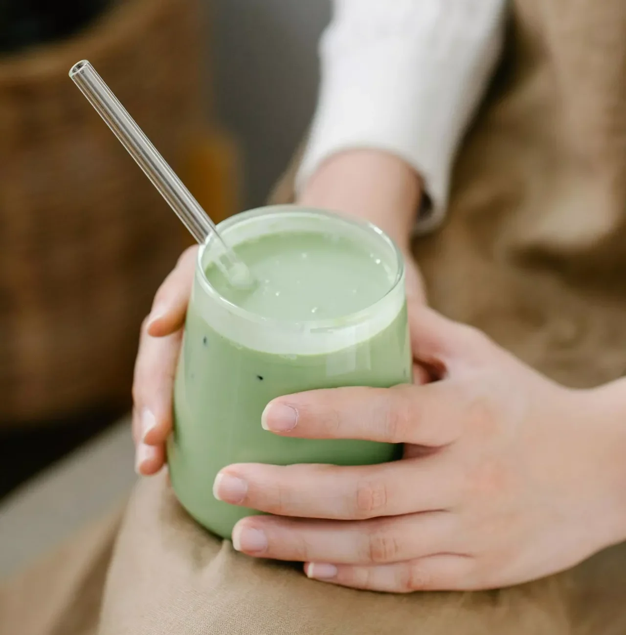 MycoVital_ Une personne tenant un verre de matcha latte vert avec une paille en verre transparent, posé sur ses genoux. La personne porte un tablier beige et un haut blanc à manches longues.