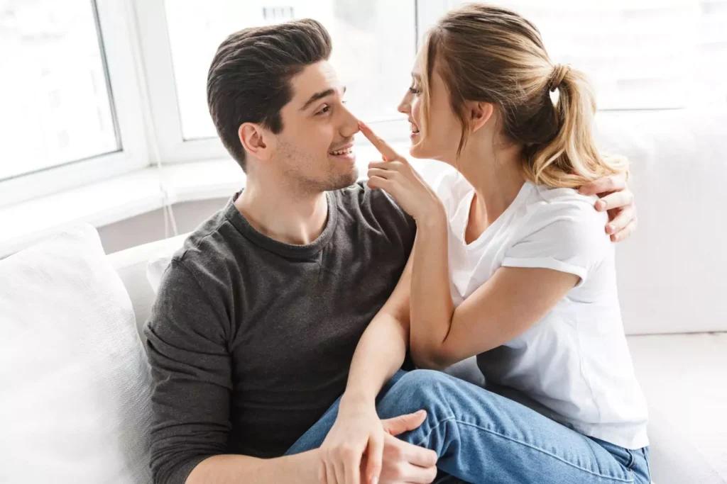 MycoVital_ A young couple sits close together on a couch, smiling warmly. The woman playfully touches the mans nose with her finger. Both appear happy and relaxed in a bright, cozy room.