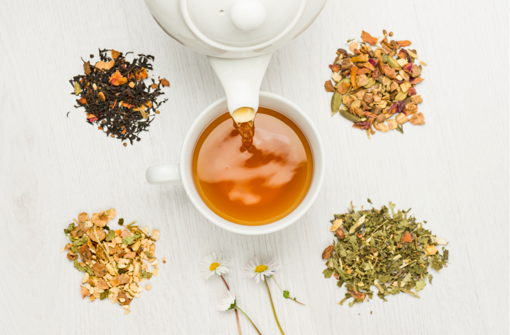 MycoVital_ Una tetera blanca vierte el té en una taza rodeada de cuatro montones de diferentes infusiones de hojas sueltas y dos flores de manzanilla, todo dispuesto sobre una superficie de madera clara.