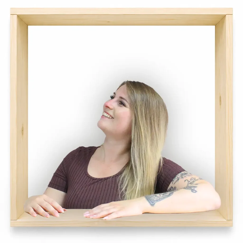 MycoVital_ Eine Frau mit langen blonden Haaren und einem Tattoo auf dem rechten Arm sitzt in einem quadratischen Holzrahmen, l&auml;chelt und blickt vor einem schlichten wei&szlig;en Hintergrund nach links oben.