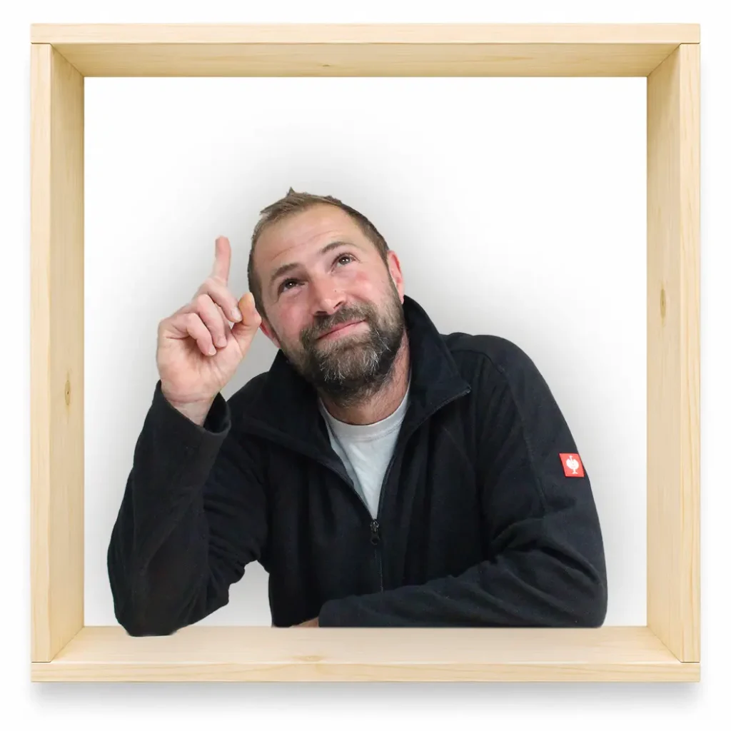 MycoVital_ Ein b&auml;rtiger Mann in einer schwarzen Jacke sitzt in einem quadratischen Holzrahmen, blickt nach oben und l&auml;chelt, w&auml;hrend er mit der rechten Hand den Zeigefinger nach oben streckt &ndash; vor einem wei&szlig;en Hintergrund.