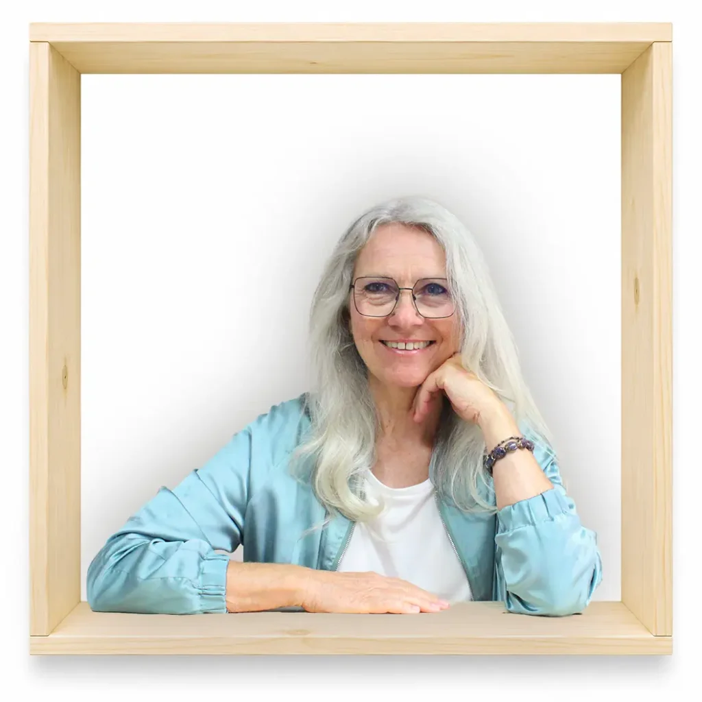 MycoVital_ Eine &auml;ltere Frau mit langen grauen Haaren und Brille l&auml;chelt, w&auml;hrend sie ihr Kinn auf eine Hand st&uuml;tzt. Sie tr&auml;gt eine hellblaue Jacke und sitzt in einem Holzrahmen vor einem wei&szlig;en Hintergrund.