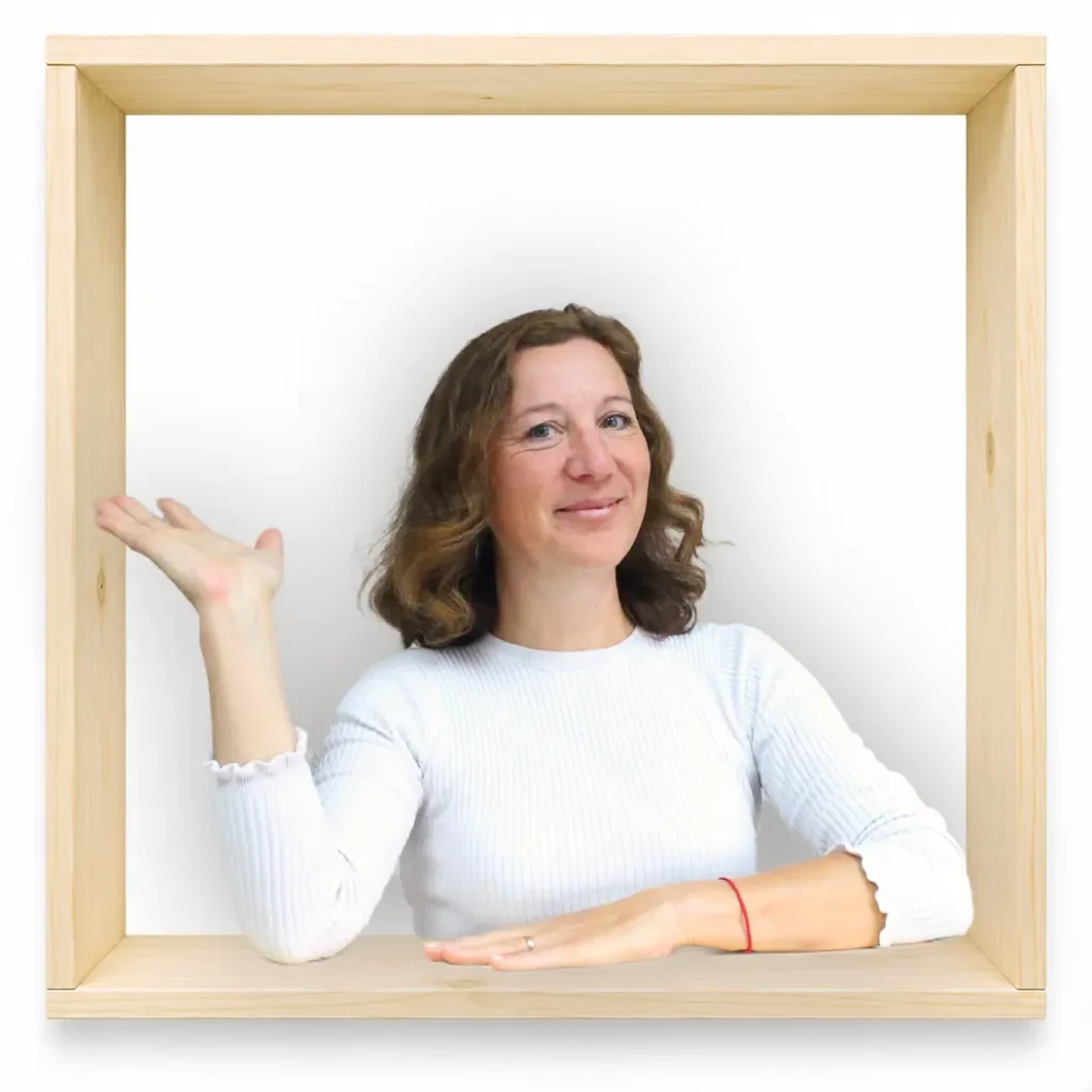 MycoVital_ Eine Frau mit welligem braunem Haar, die ein wei&szlig;es Langarmhemd tr&auml;gt, sitzt in einem quadratischen Rahmen aus hellem Holz. Sie l&auml;chelt und hebt ihre linke Hand in einer einladenden Geste, w&auml;hrend ihre rechte Hand auf dem Rahmen ruht.