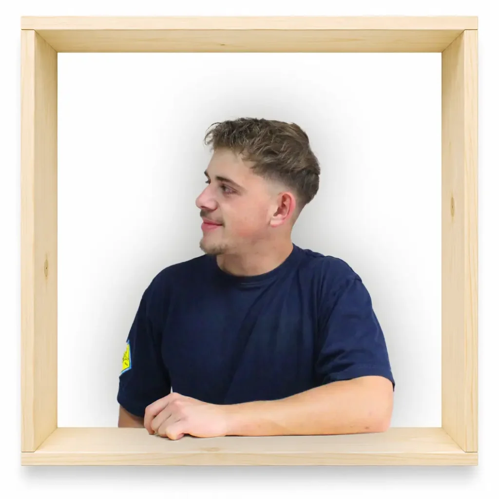 MycoVital_ Ein junger Mann mit kurzen braunen Haaren, der ein marineblaues T-Shirt tr&auml;gt, steht in einem gro&szlig;en, leeren Holzrahmen vor einem wei&szlig;en Hintergrund, blickt nach links und l&auml;chelt.