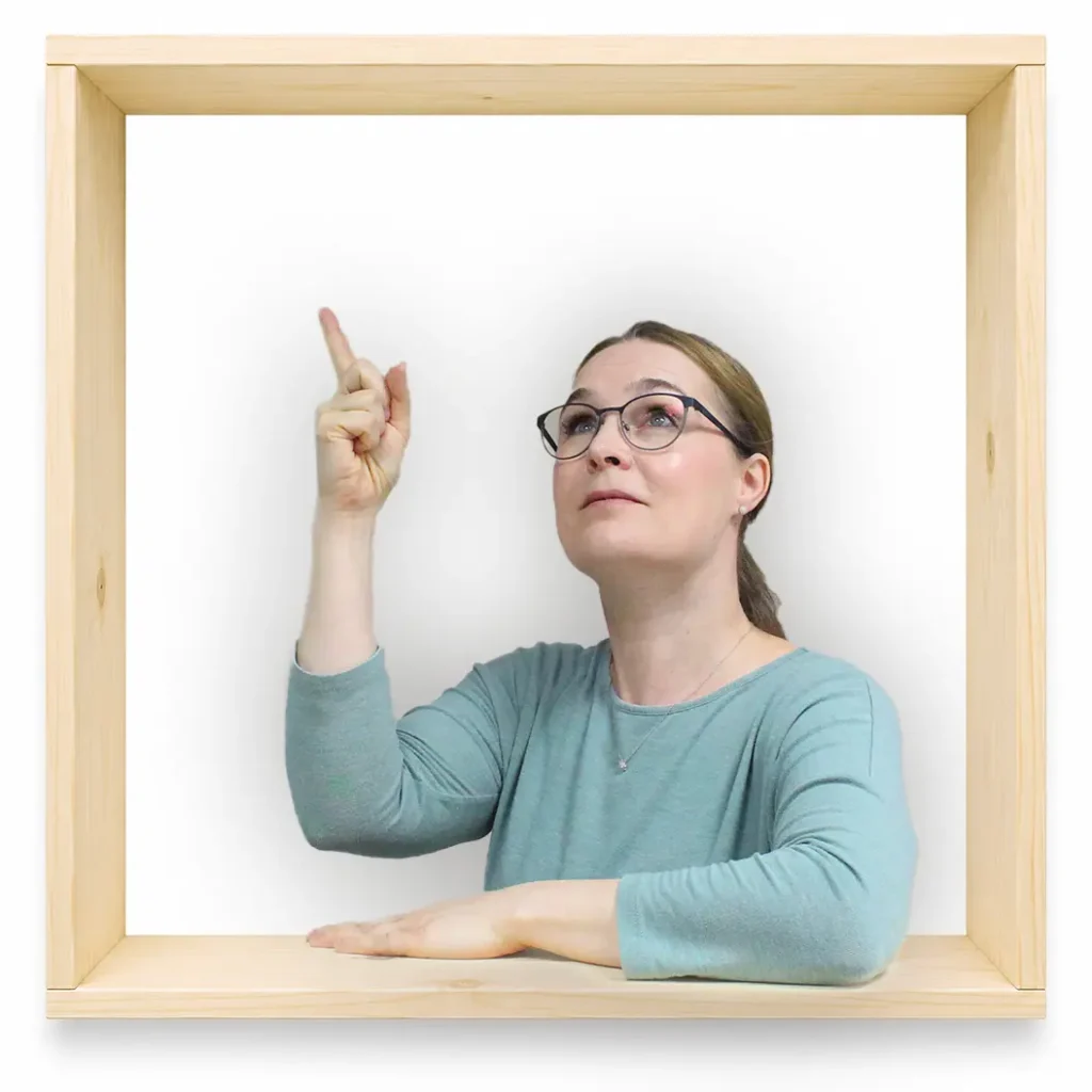 MycoVital_ Eine Frau mit Brille und einem hellblauen Oberteil sitzt in einem Holzrahmen, blickt nach oben und zeigt mit ihrem rechten Zeigefinger nach oben. Der Hintergrund ist schlicht wei&szlig;.