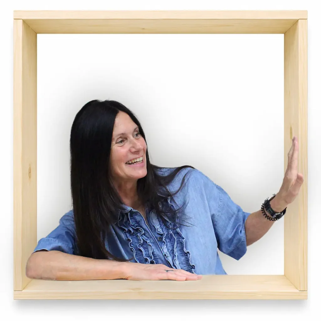MycoVital_ Eine Frau mit langen dunklen Haaren, die ein blaues R&uuml;schenhemd tr&auml;gt, l&auml;chelt und blickt zur Seite, w&auml;hrend sie sich auf die Ecke eines gro&szlig;en Holzrahmens vor einem schlichten wei&szlig;en Hintergrund st&uuml;tzt und diese ber&uuml;hrt.
