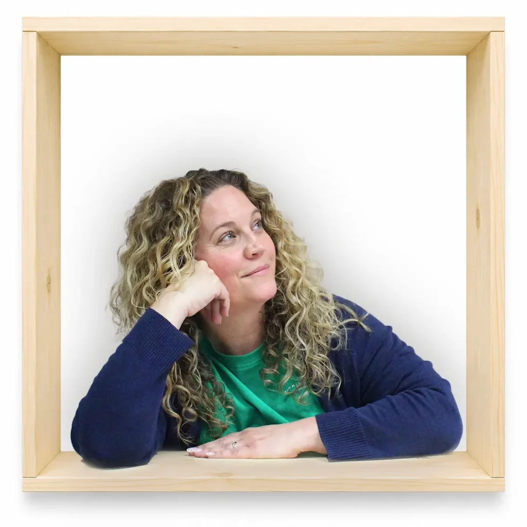MycoVital_ Eine Frau mit lockigem blondem Haar, die ein gr&uuml;nes Shirt und einen marineblauen Pullover tr&auml;gt, st&uuml;tzt ihren Kopf auf die Hand und blickt nach oben, w&auml;hrend sie in einem quadratischen Holzrahmen vor einem wei&szlig;en Hintergrund sitzt.